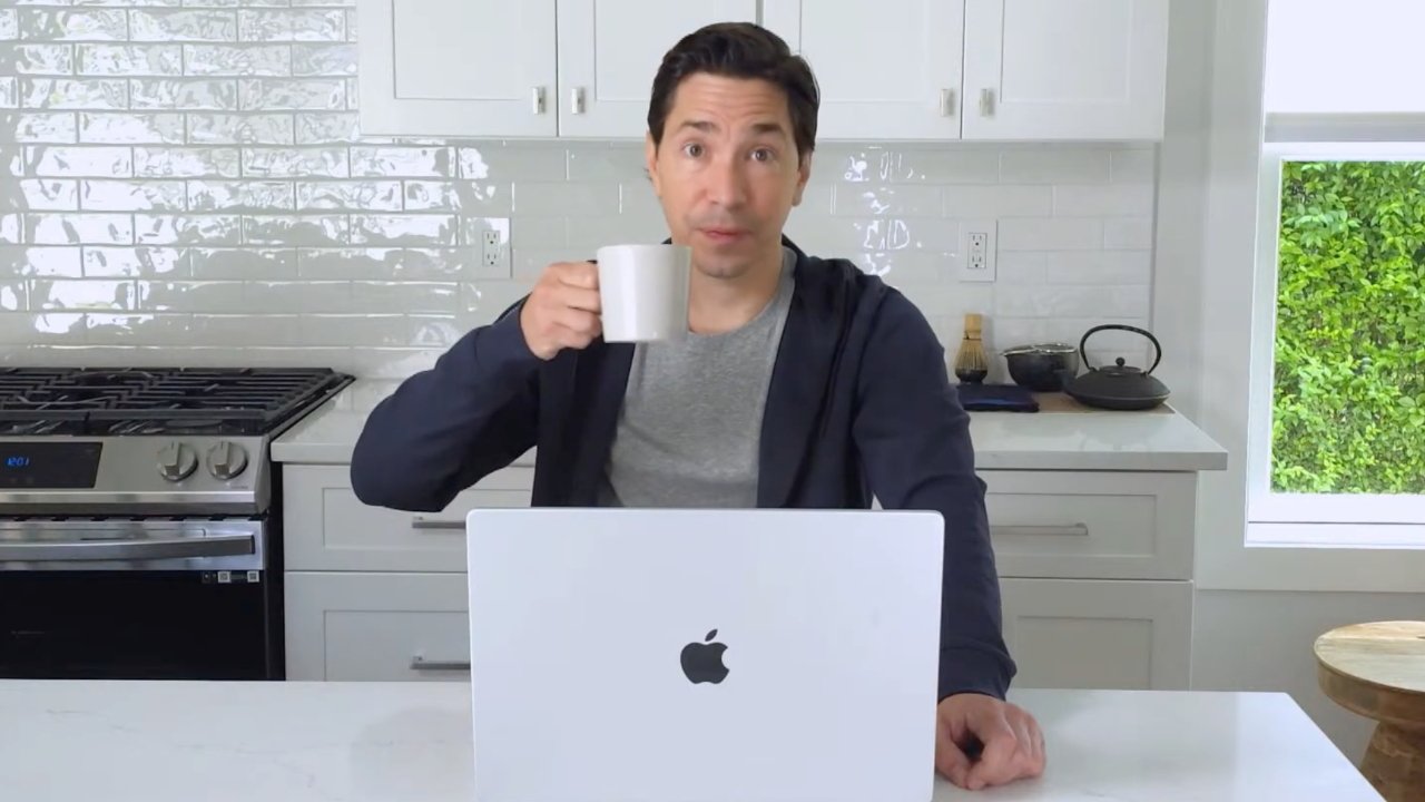 Person sitting in a white kitchen, holding a mug, with an open laptop on the counter and a stove in the background.