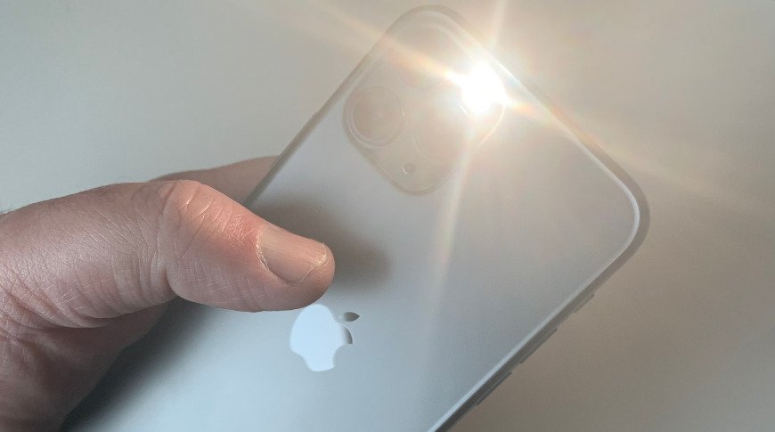 Hand holding silver iPhone with camera flash activated, emitting a bright light against a white background.