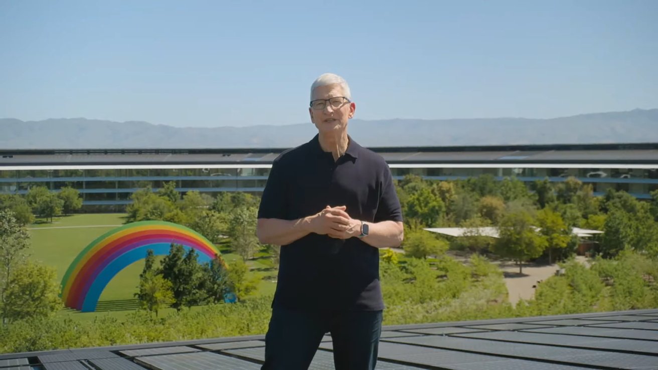Tim Cook standing outside with hands clasped, wearing glasses, in front of modern building and colorful rainbow arch among green trees, with mountains in the background.
