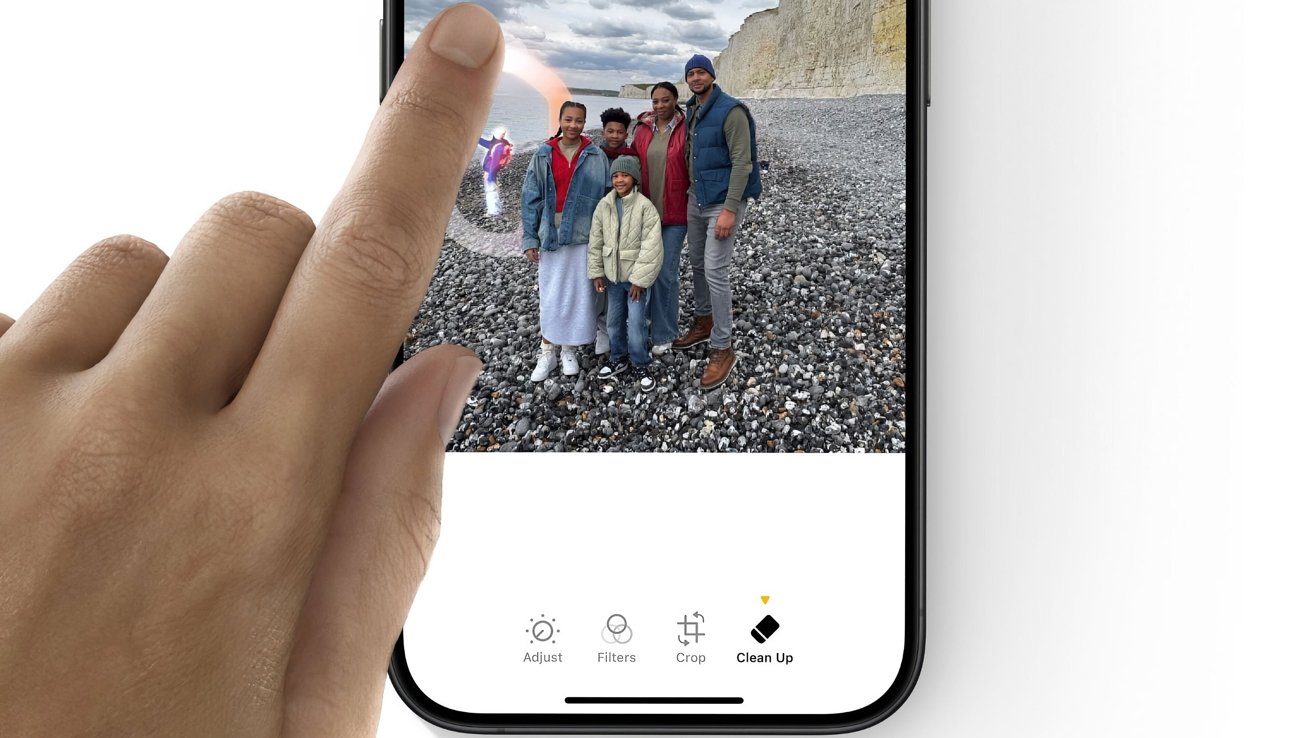 A hand touches a smartphone screen, editing a family photo on a rocky beach using the 'Clean Up' tool.