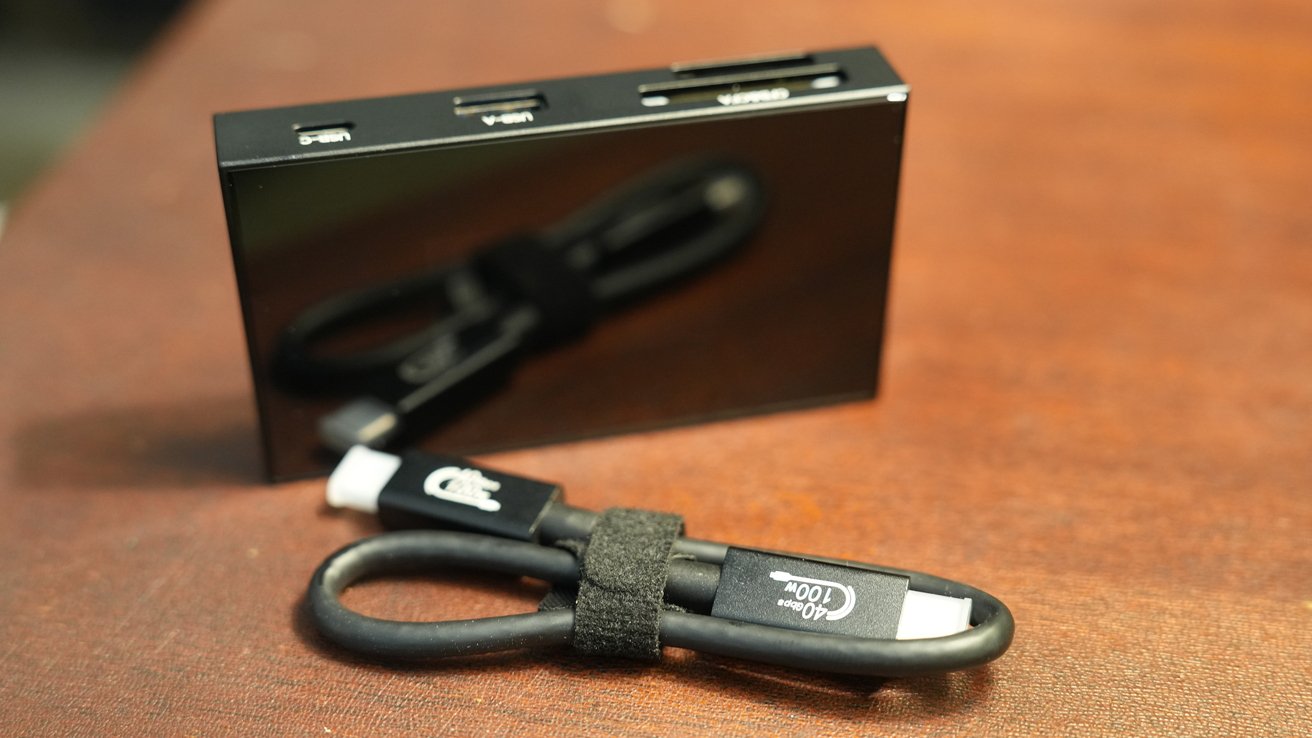 Black multi-port adapter with USB-C, USB-A, and SD card slots on a brown surface, next to a coiled black charging cable.