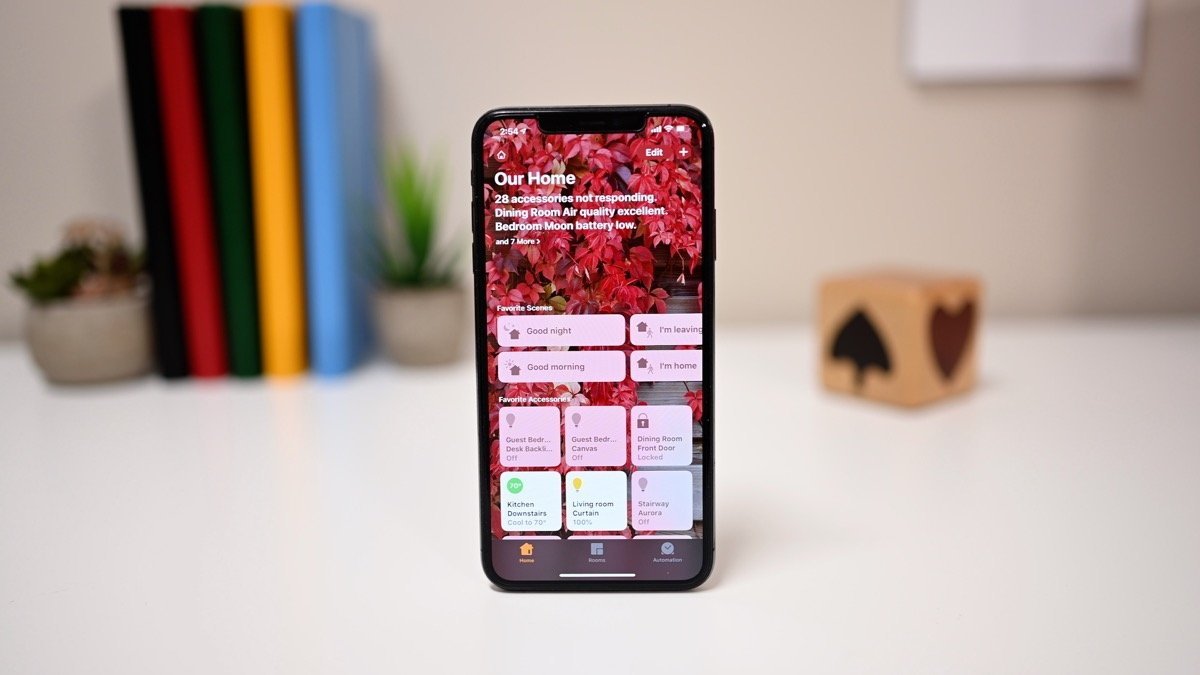 Smartphone showing a home automation app with various controls. Blurred background includes colorful books, small potted plants, and a dice block.