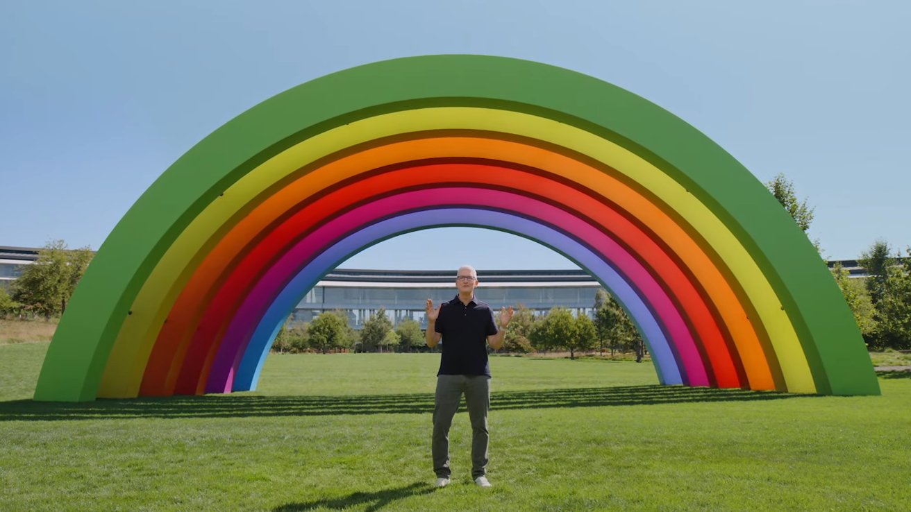 Man standing under large, colorful rainbow sculpture on bright green grass, with a modern building and clear blue sky in the background.