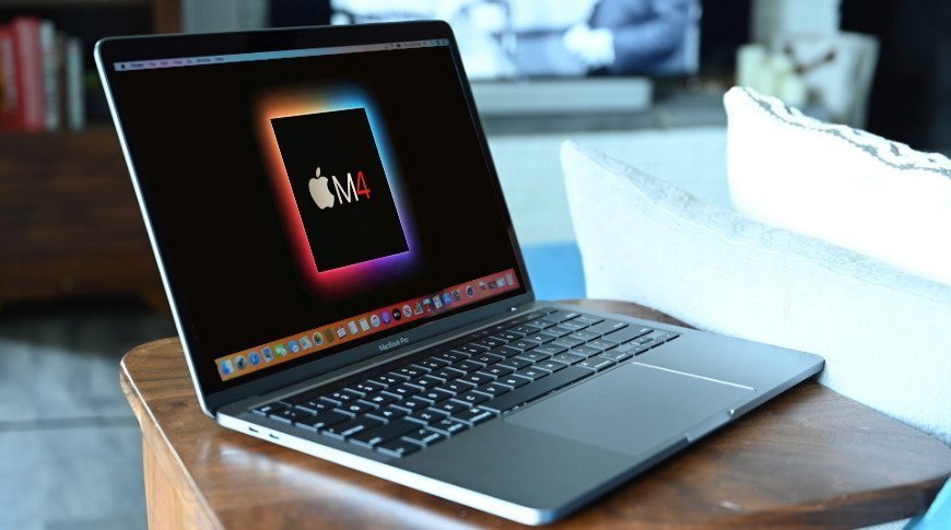 A MacBook Pro on a wooden table displaying a colorful Apple M4 logo on its screen, with blurred background furniture and bookshelf.