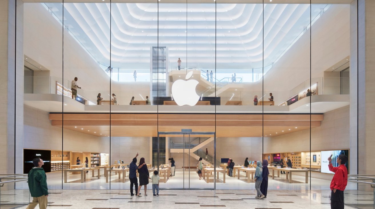 Modern two-story store with a large glass facade, featuring an illuminated apple logo, various product displays, and several customers browsing inside.