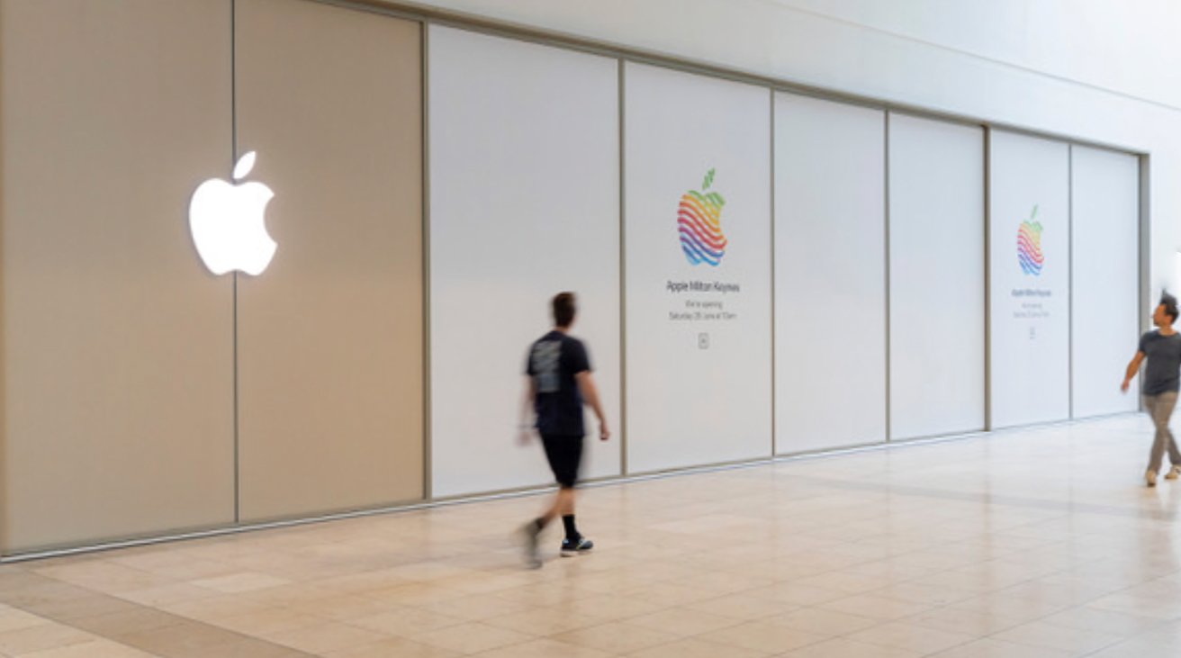 Apple Store with white logo on beige wall, colorful apple icons, text announcing new store opening, two people walking, light-toned tile floor.