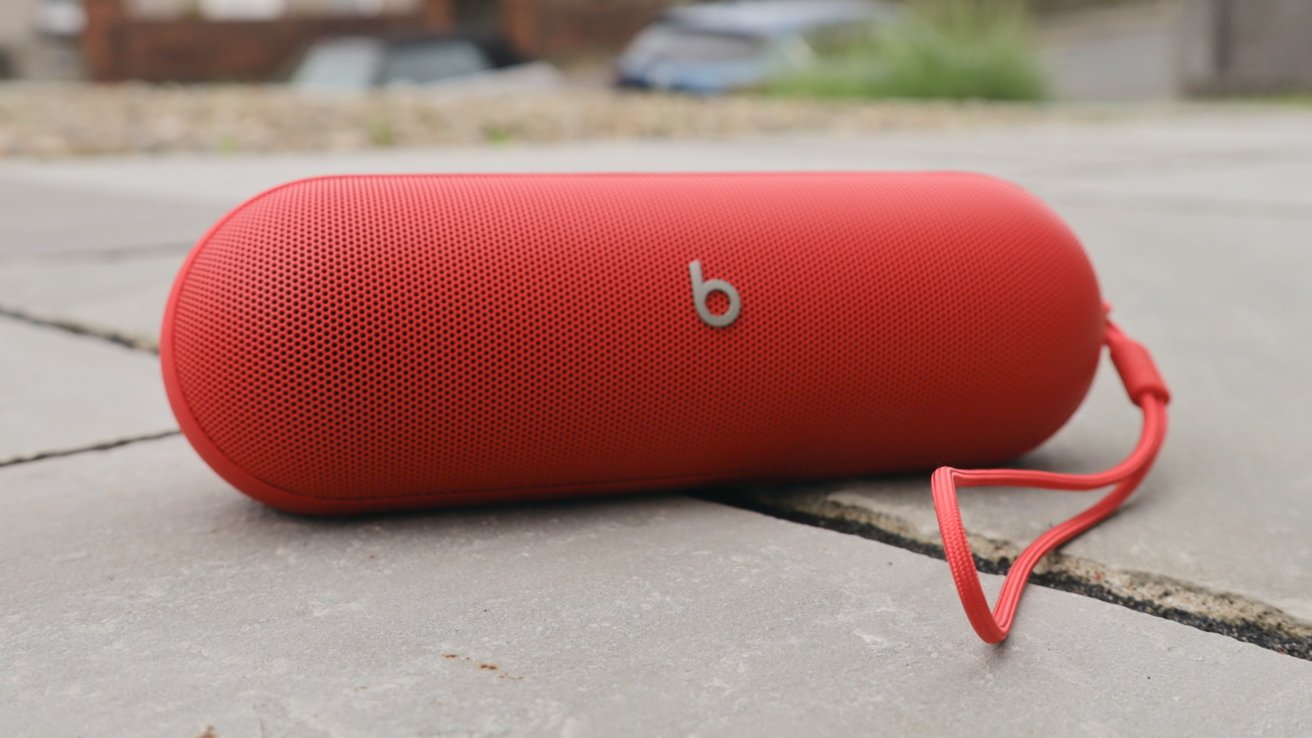 Red cylindrical portable speaker with a small letter 'b' logo, resting on a tiled surface outdoors with grass and blurred background. A red strap is attached to one end.
