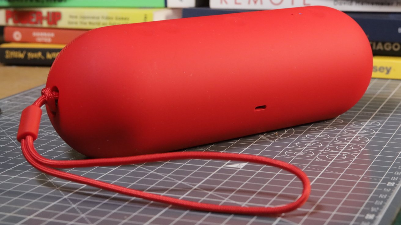 Red cylindrical portable speaker with a wrist strap, resting on a cutting mat. Books stacked in the background.