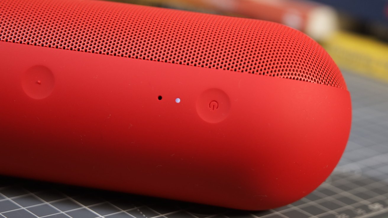 A red cylindrical speaker with mesh top, showing two small buttons and indicator lights on a gray grid-patterned surface.