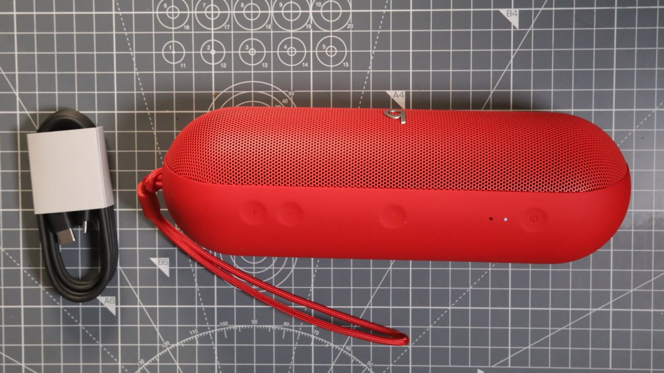 Red portable speaker with a wrist strap next to a coiled black charging cable on a cutting mat with gridlines.