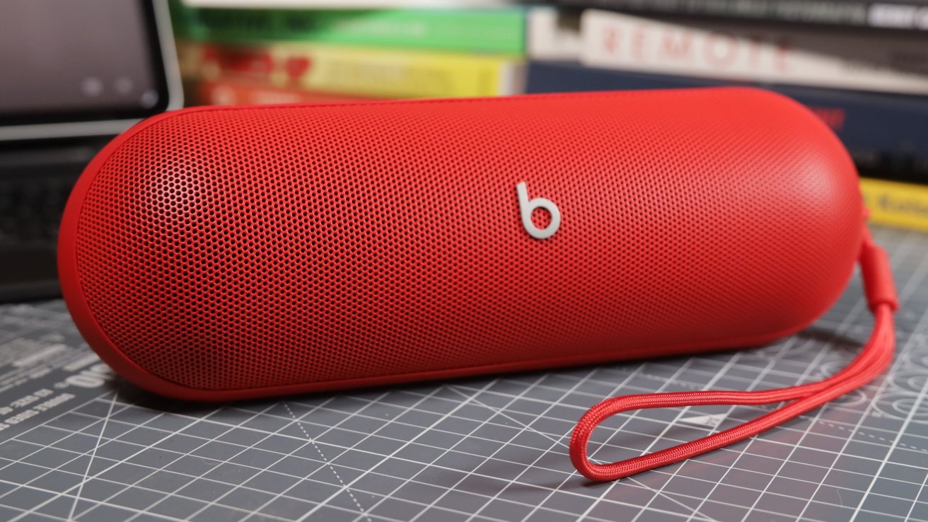 Red portable Bluetooth speaker with a perforated grille and a carrying strap, placed on a grid-patterned surface in front of a stack of books.