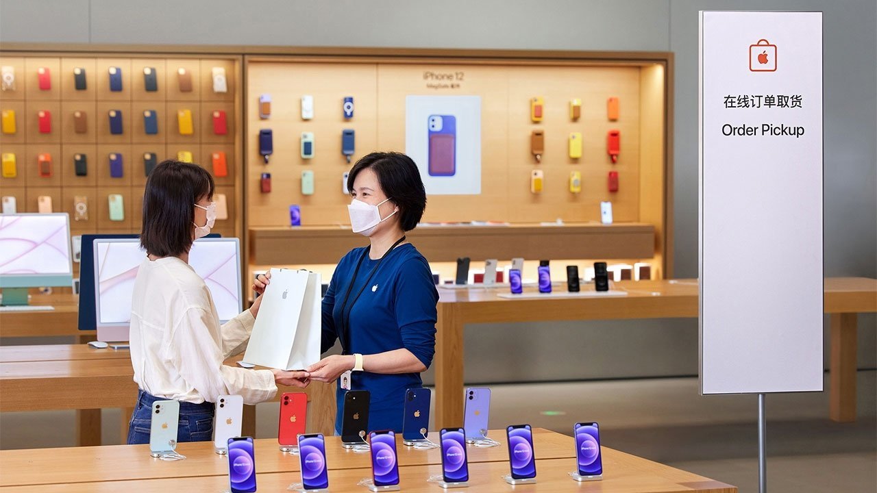 Two masked women exchange a shopping bag in an Apple store with iPhones being displayed. A sign reads 'Order Pickup' in English and another language.