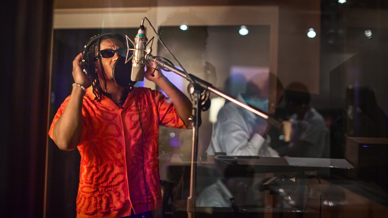 A person in a red shirt and sunglasses sings into a microphone in a recording studio.