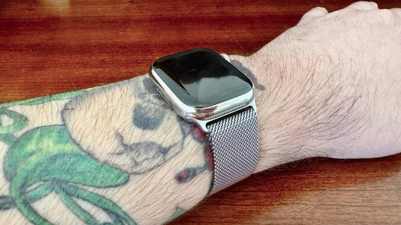 Wrist with a tattoo wearing a smartwatch with a metal mesh band, against a wooden surface.