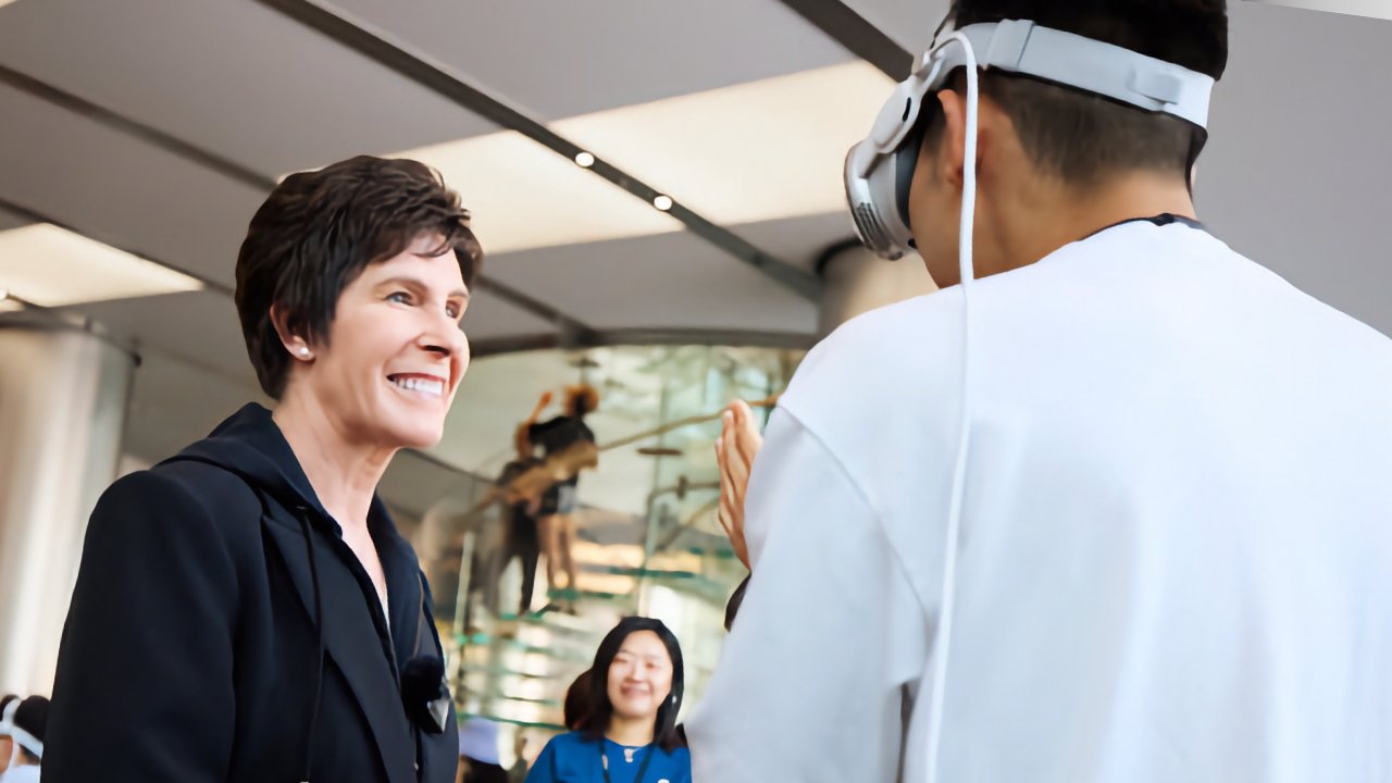 Smiling woman in a dark jacket talking to a man wearing headphones. Another person is in the background, and they are indoors.
