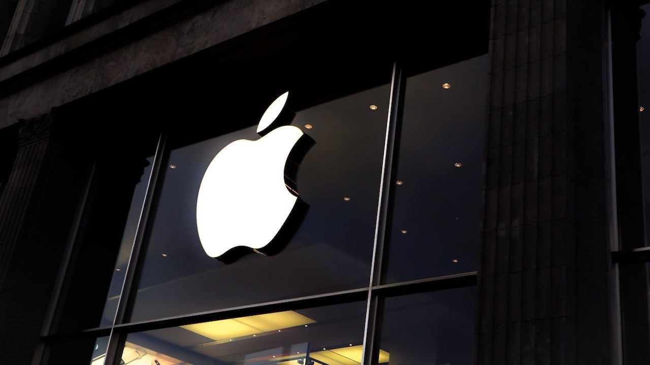 Large, glowing white Apple logo on a dark storefront window, surrounded by columns, reflecting lights from inside the store.