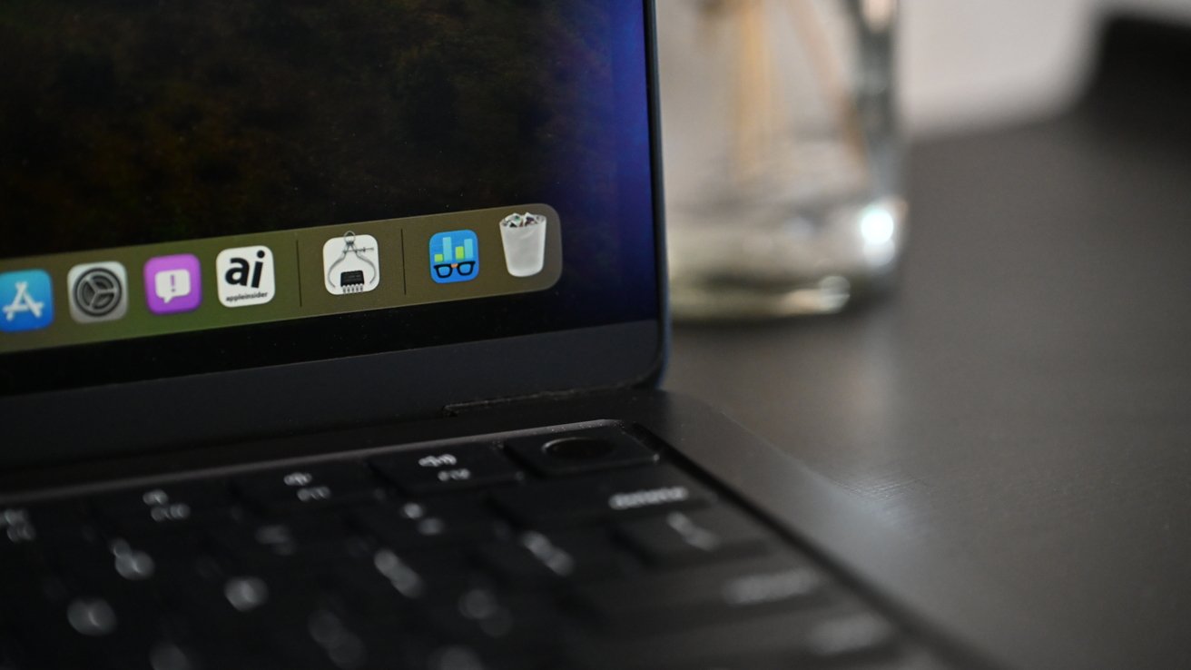 MacBook screen displaying app icons at the bottom, including App Store, System Preferences, Messages, and a trash can; keyboard and blurry background visible.