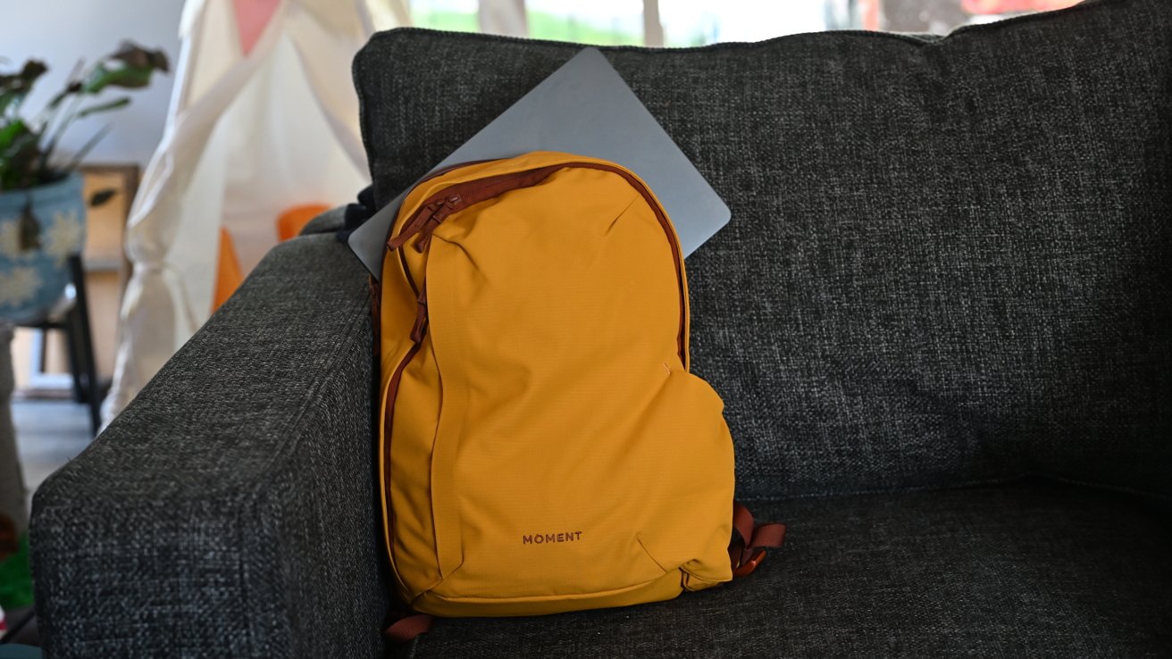 Yellow backpack on a dark gray couch with a closed laptop partly underneath, indoor plant in the background.