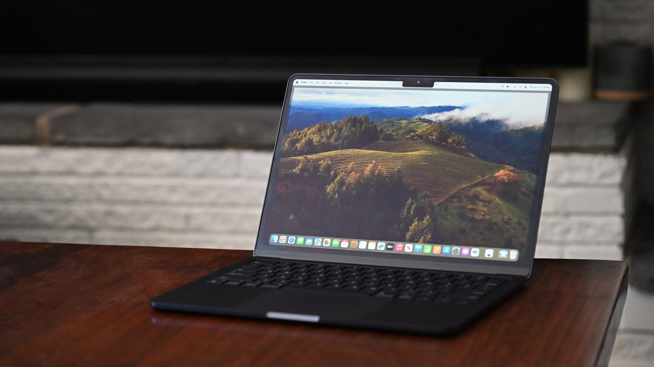 Laptop computer on a wooden table with a scenic landscape on the screen, showing hills and trees. Background of a gray fireplace.