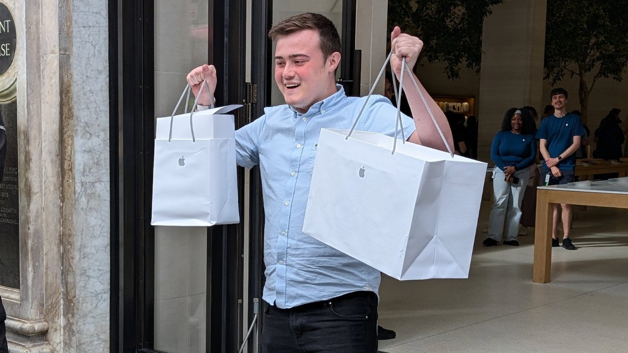 Man holding large Apple-branded shopping bags, standing inside a store with people in the background.