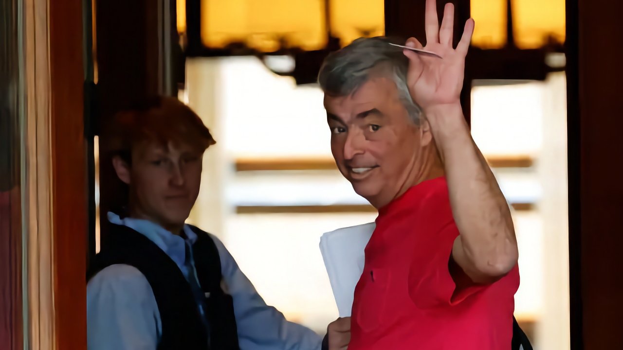 Smiling man in a red shirt waves while holding a document, standing next to a person in the background with a vest and light blue shirt.