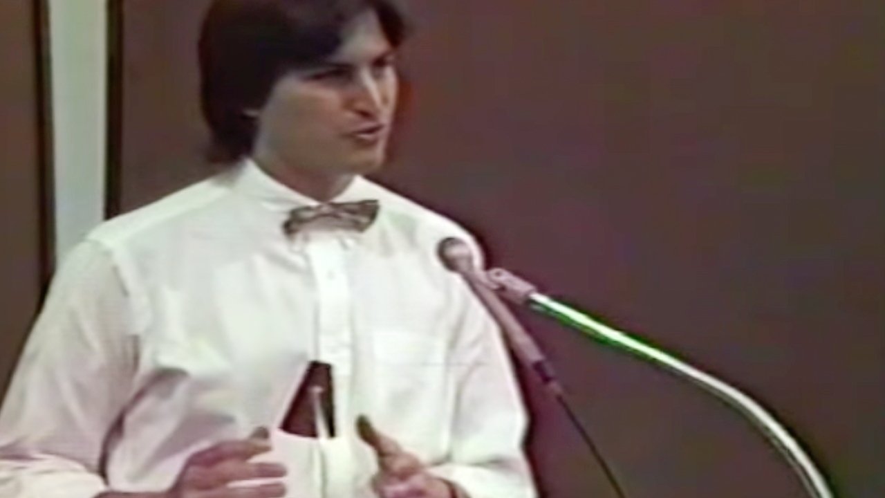 Man in a white shirt and bow tie speaking at a microphone with a brown background.