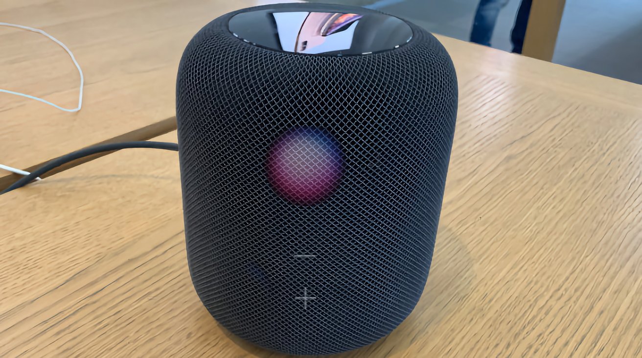 Smart speaker with a mesh exterior, displaying a colorful dot on the front, placed on a wooden table.