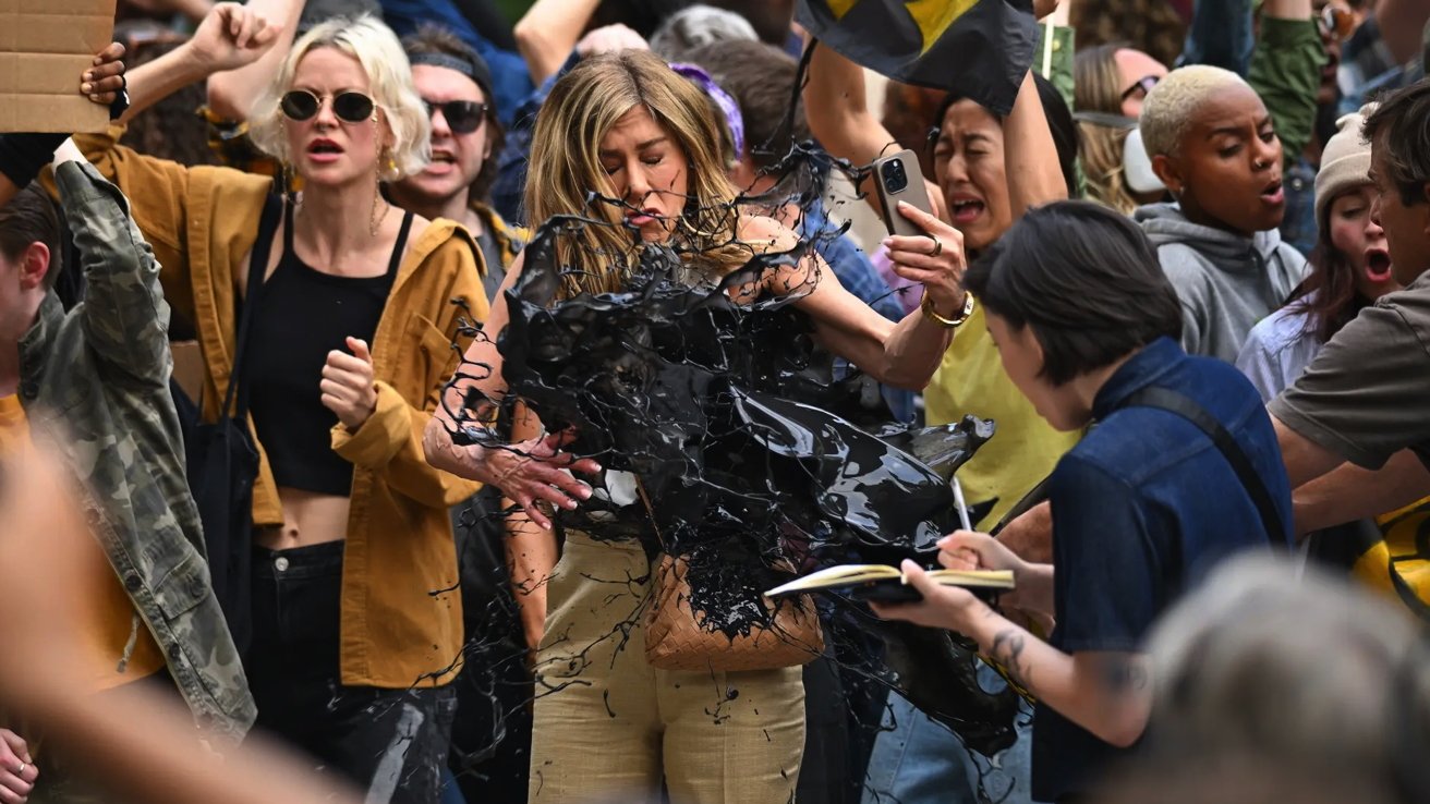 A protesting crowd with a woman covered in black paint, others holding signs and smartphones in the background.