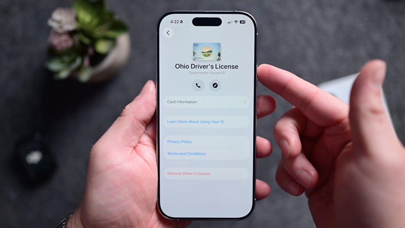 Hands holding a smartphone displaying an Ohio Driver's License app interface with user options and menu buttons; background shows blurred greenery.
