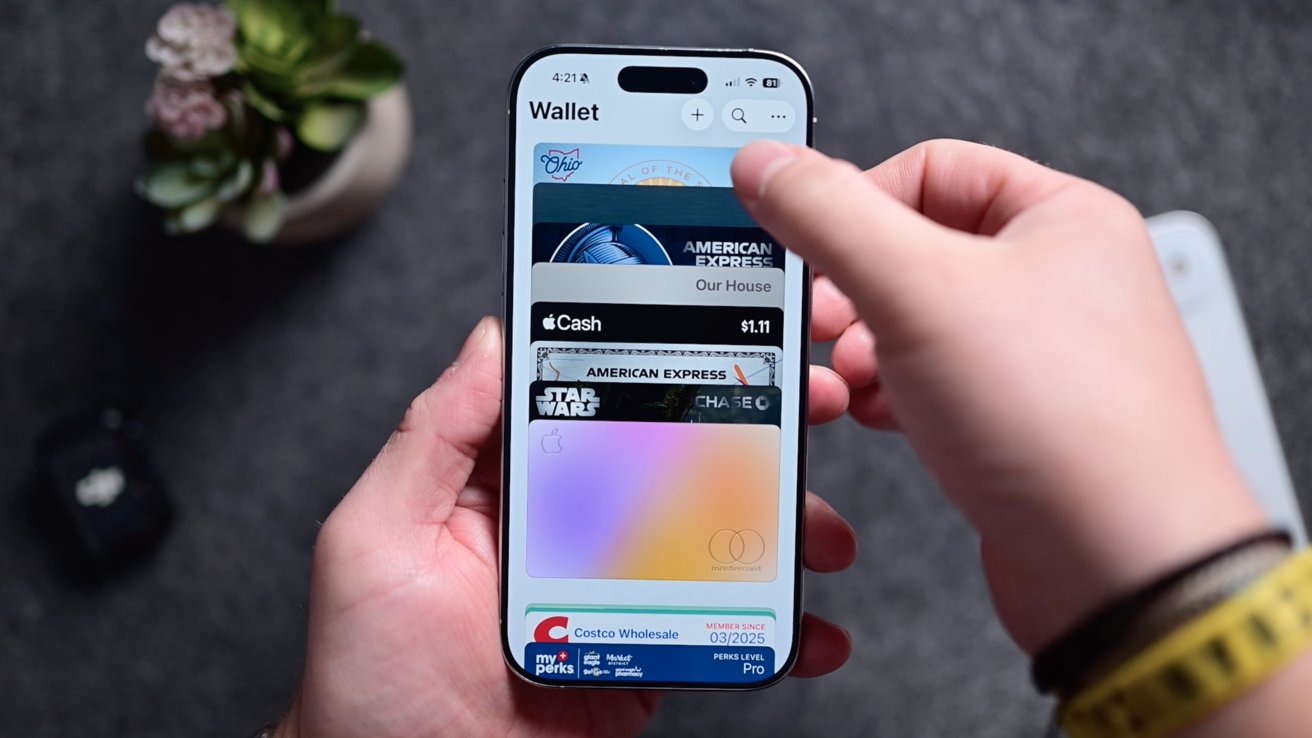 A person holds a smartphone displaying a digital wallet with several cards visible, including American Express, Apple Cash, and Costco, on a textured gray background.