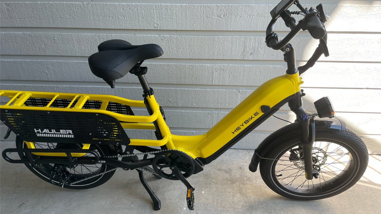 A yellow electric cargo bike with a black rear rack, dual passenger seat, and large tires, leaning against a light gray wooden wall.