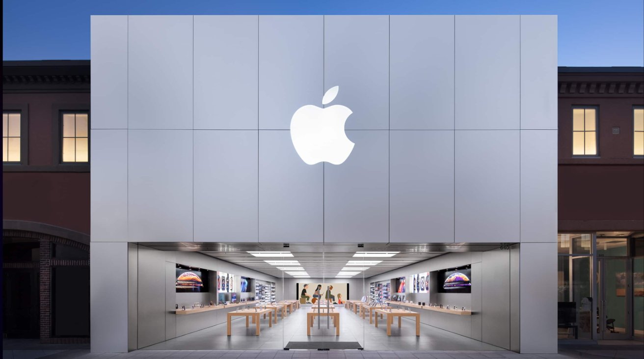 Apple Store exterior with a large Apple logo above the entrance. Inside are tables with products and large advertising screens on the walls.