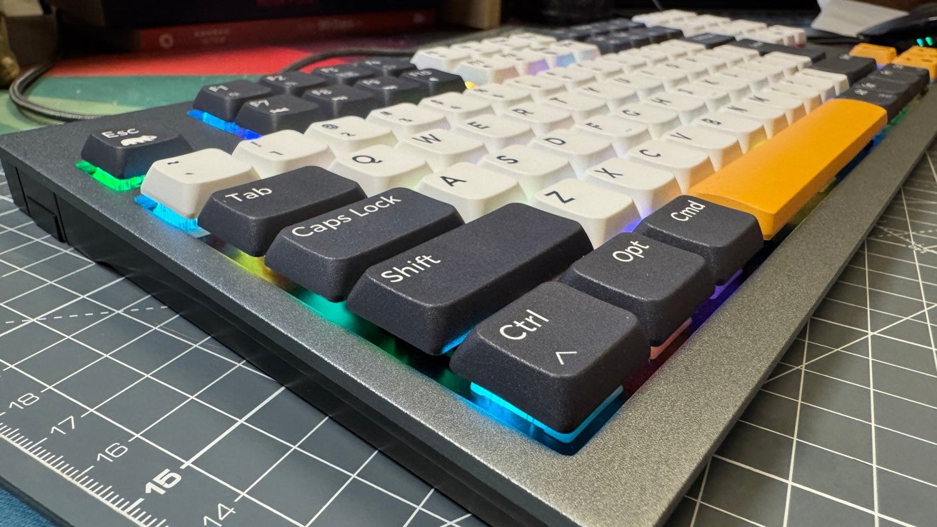 Mechanical keyboard with colorful backlighting, black and white keys, and an orange spacebar, placed on a grid-patterned desk mat.