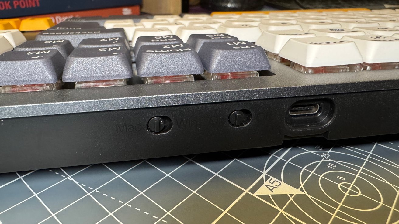 Close-up of a mechanical keyboard with grey and white keycaps, a USB-C port, and switches visible. Background includes a cutting mat and a stack of books.