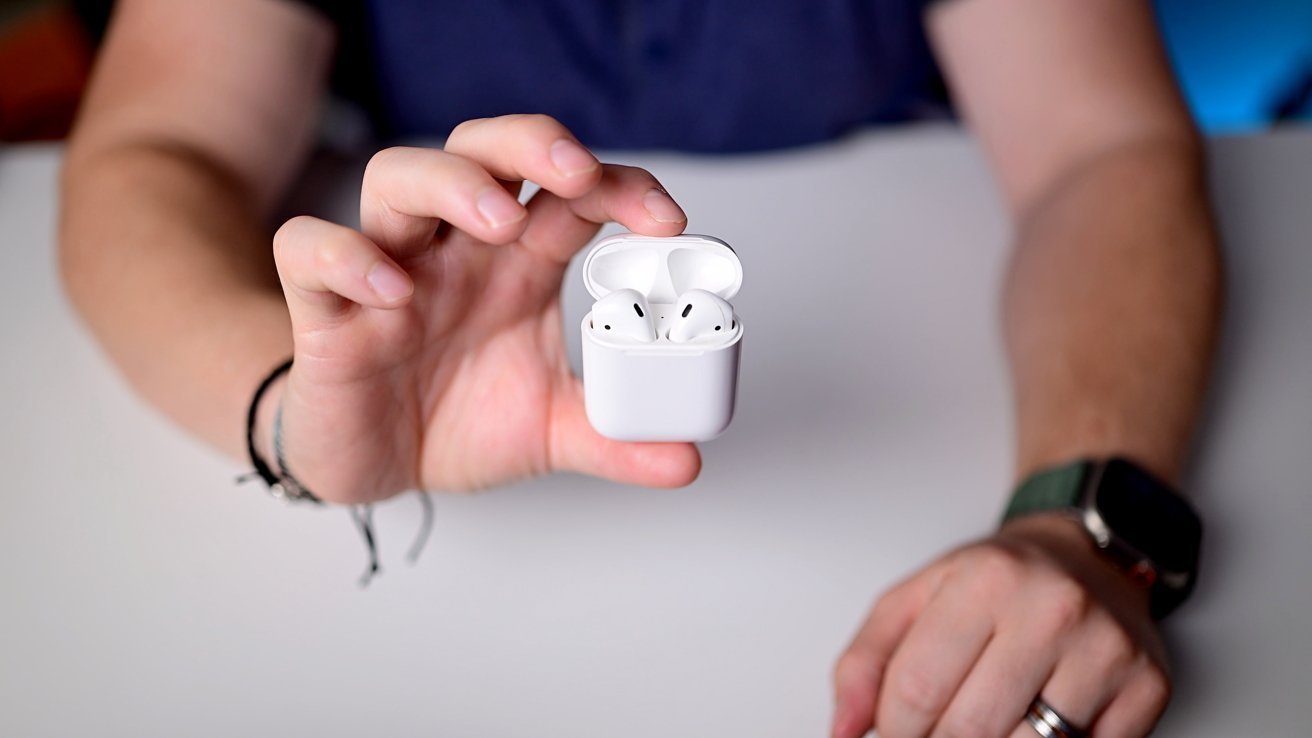 Hand holding an open white case with wireless earbuds on a white table. Wristwatch and bracelets are visible on the person's wrist.