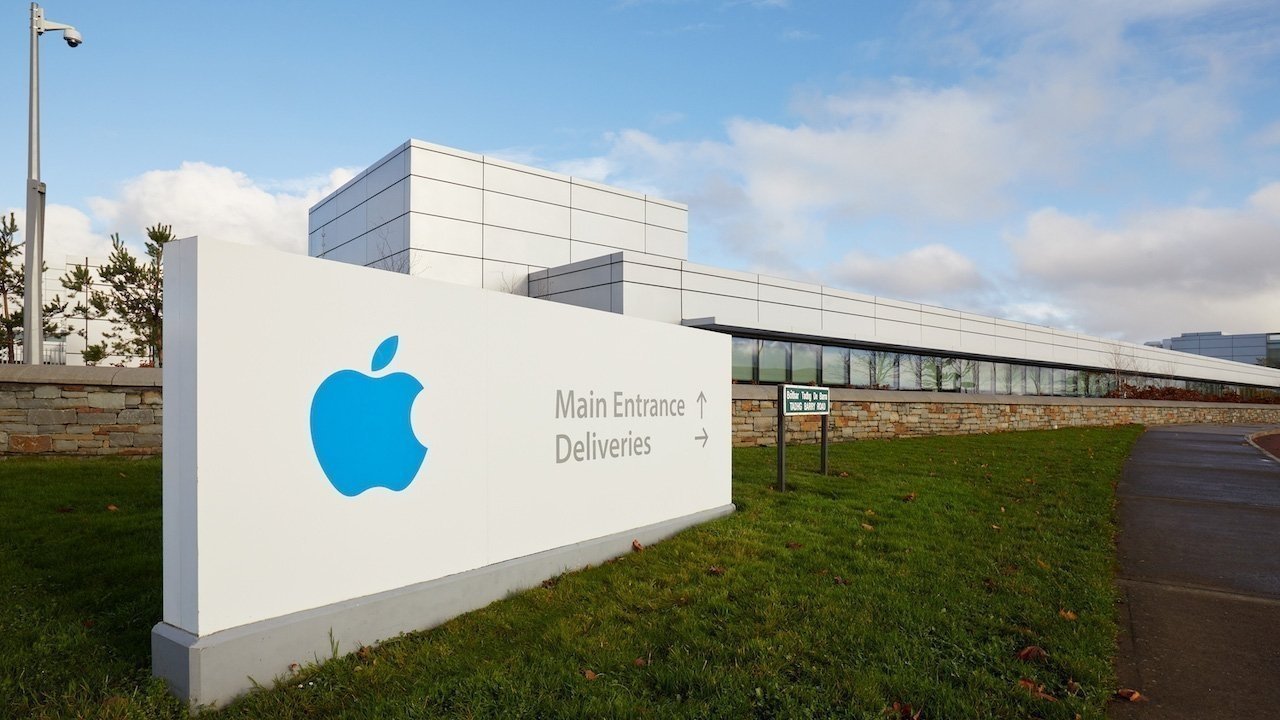 White building with a large blue Apple logo on a sign, indicating directions for Main Entrance and Deliveries. The background includes trees and a partly cloudy sky.