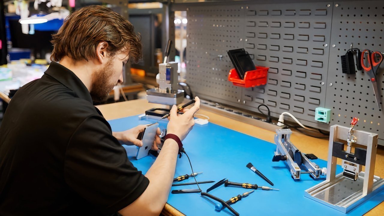 Person repairing an iPhone at a workbench with tools and equipment, blue mat, and pegboard with hanging items in a workshop setting.