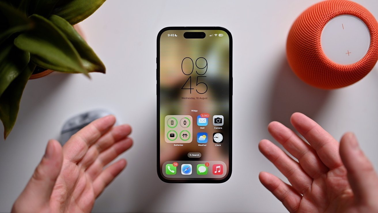 Smartphone floating between hands, surrounded by a potted plant and an orange speaker on a white surface. The phone screen displays time and various app icons.