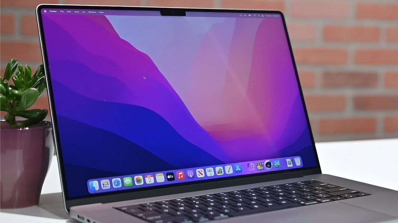 Laptop with a colorful screen displaying a purple and pink gradient wallpaper. Potted plant on the left and a brick wall background.