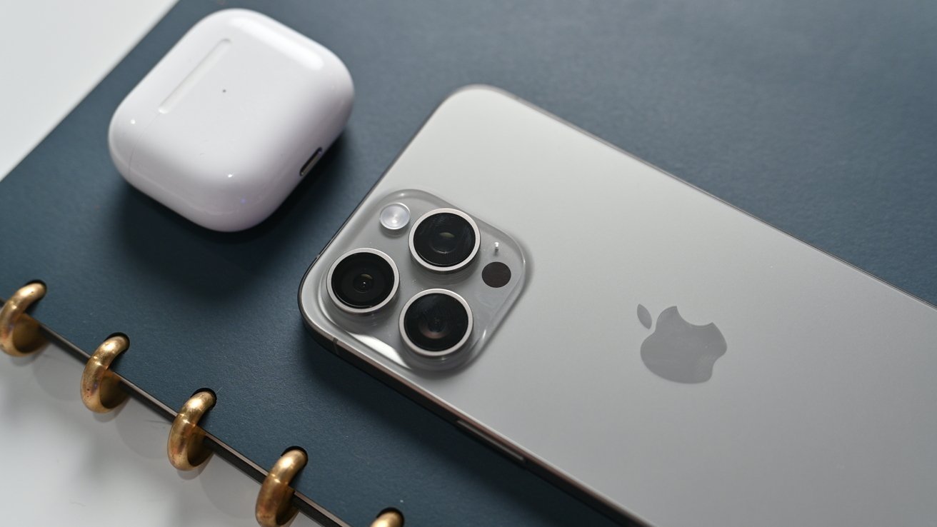 Silver iPhone with triple cameras and white AirPods case on a dark gray notebook with golden spiral binding.