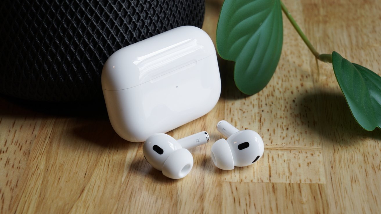 White wireless earbuds and charging case on a wooden surface with a green leaf and black mesh object in the background.