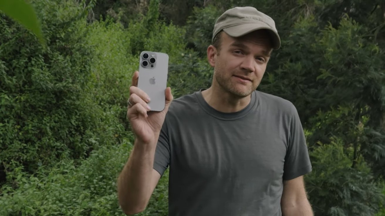 Man wearing a gray shirt and cap holds up a smartphone in front of lush greenery.