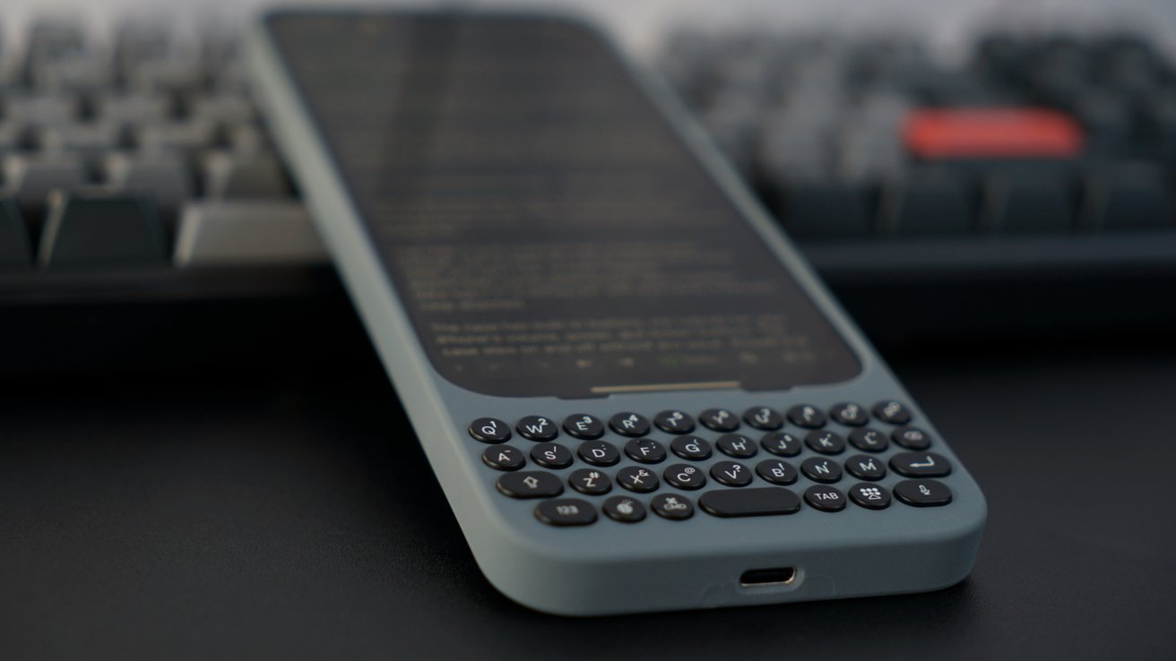 Black smartphone with physical keyboard case lying on a desk in front of a computer keyboard.