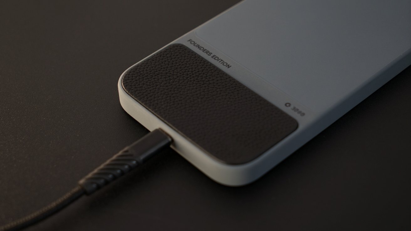 Close-up of a smartphone back labeled 'Founders Edition' with a textured black panel, connected to a charging cable.