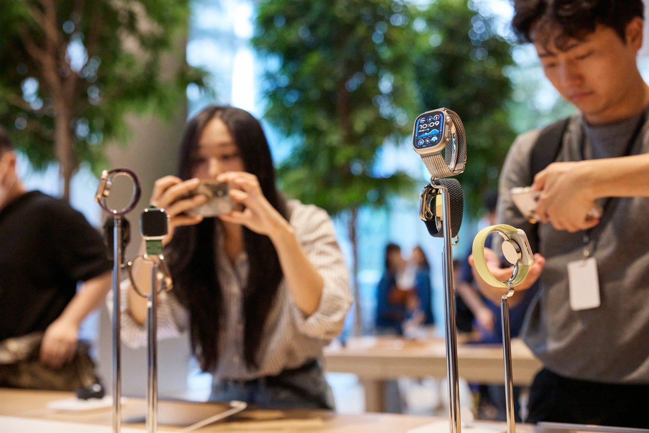 People photographing smartwatches displayed on stands in a store with trees in the background.