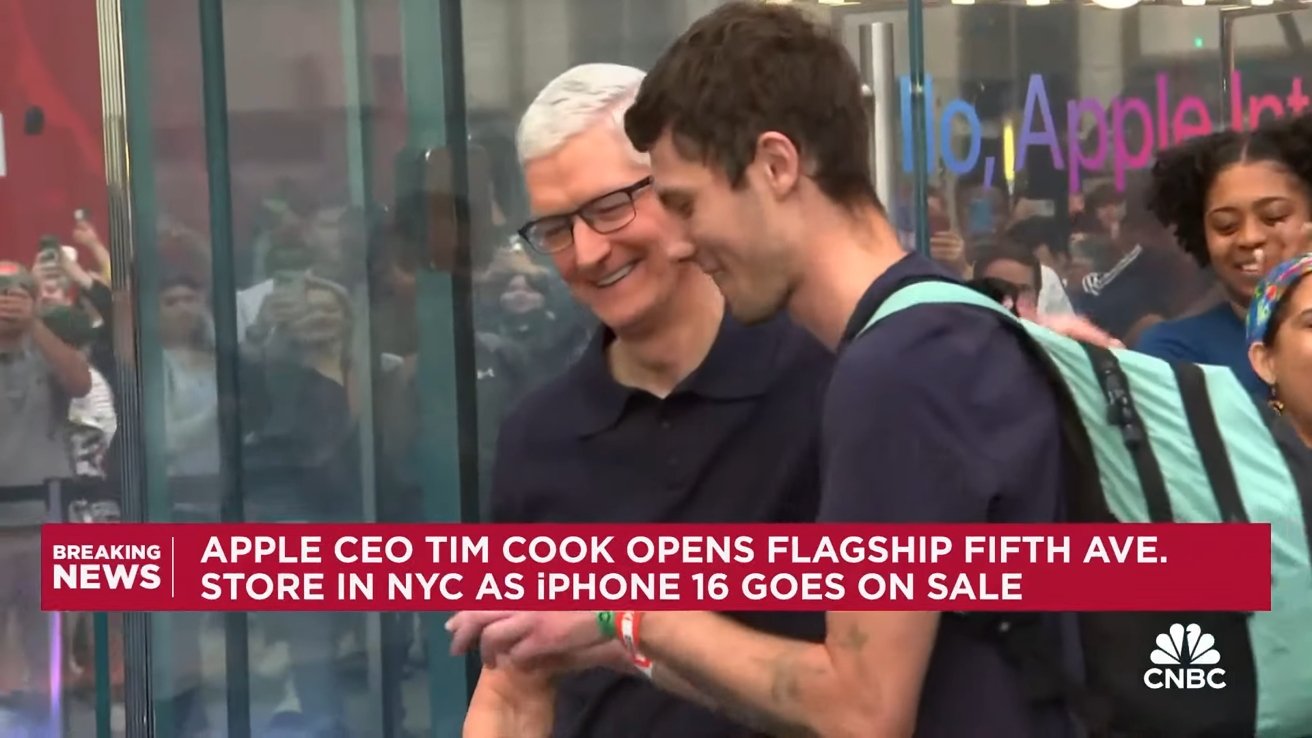 Apple CEO smiling with a customer inside the new flagship Fifth Ave. store in NYC, surrounded by people taking photos.