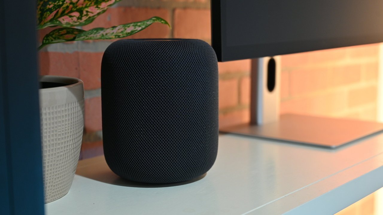 First-gen HomePod on a white desk next to a potted plant and a computer monitor with a brick wall background.