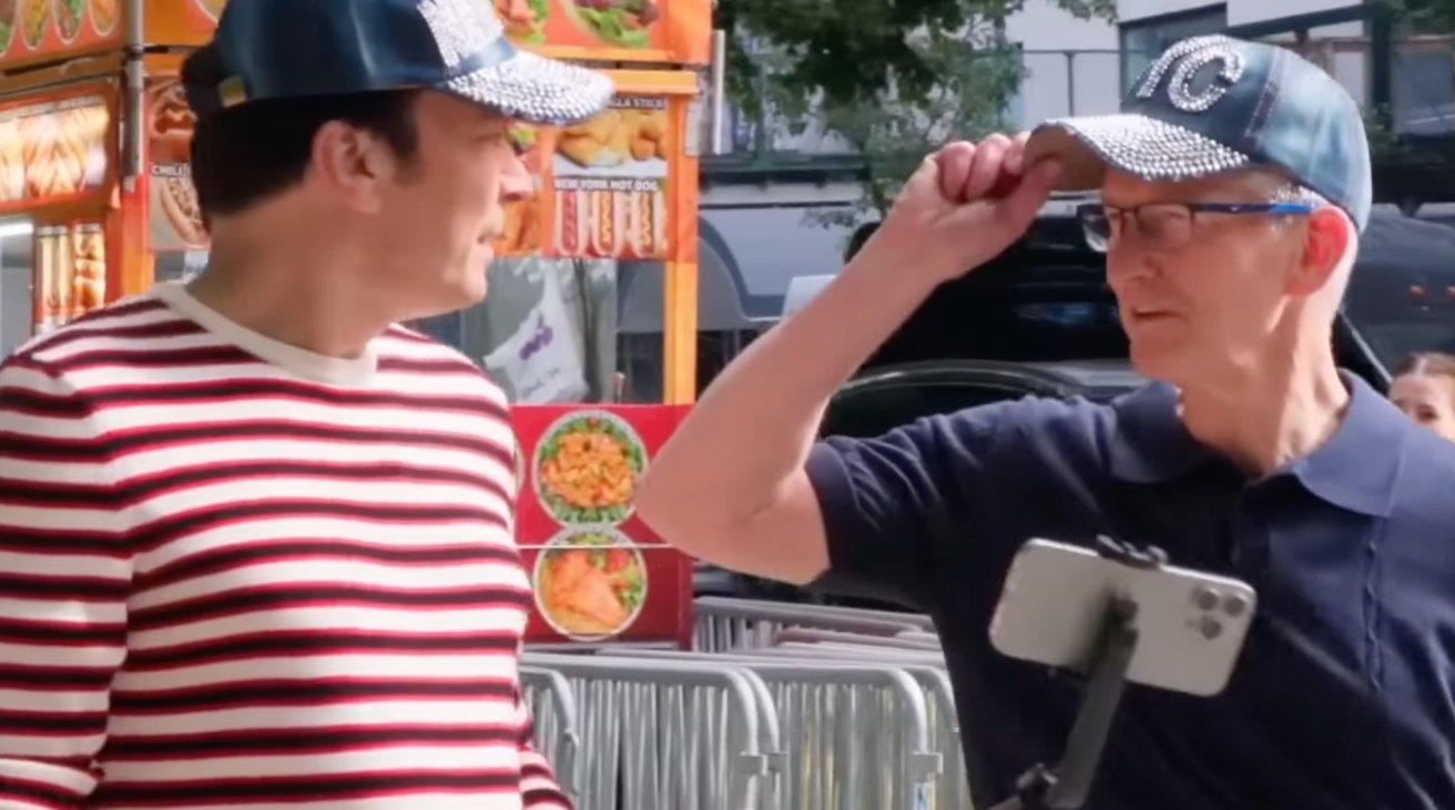 Two men in hats conversing near a food cart, one wearing a striped sweater, the other in a solid-colored shirt holding a phone on a selfie stick.