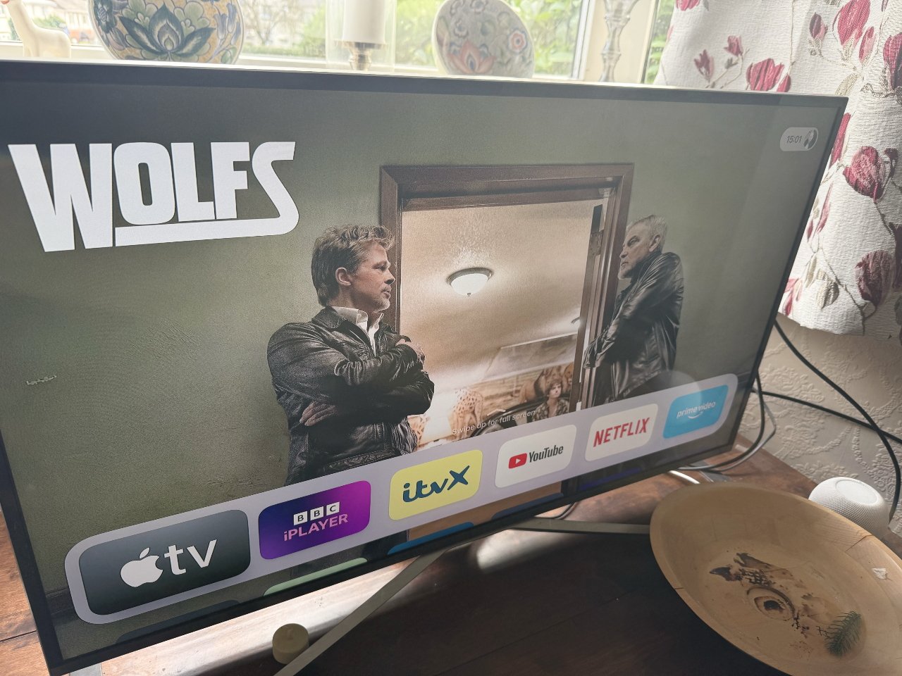 TV screen displaying a show called 'Wolfs' featuring two men in leather jackets, with streaming service icons below and a wooden bowl nearby.