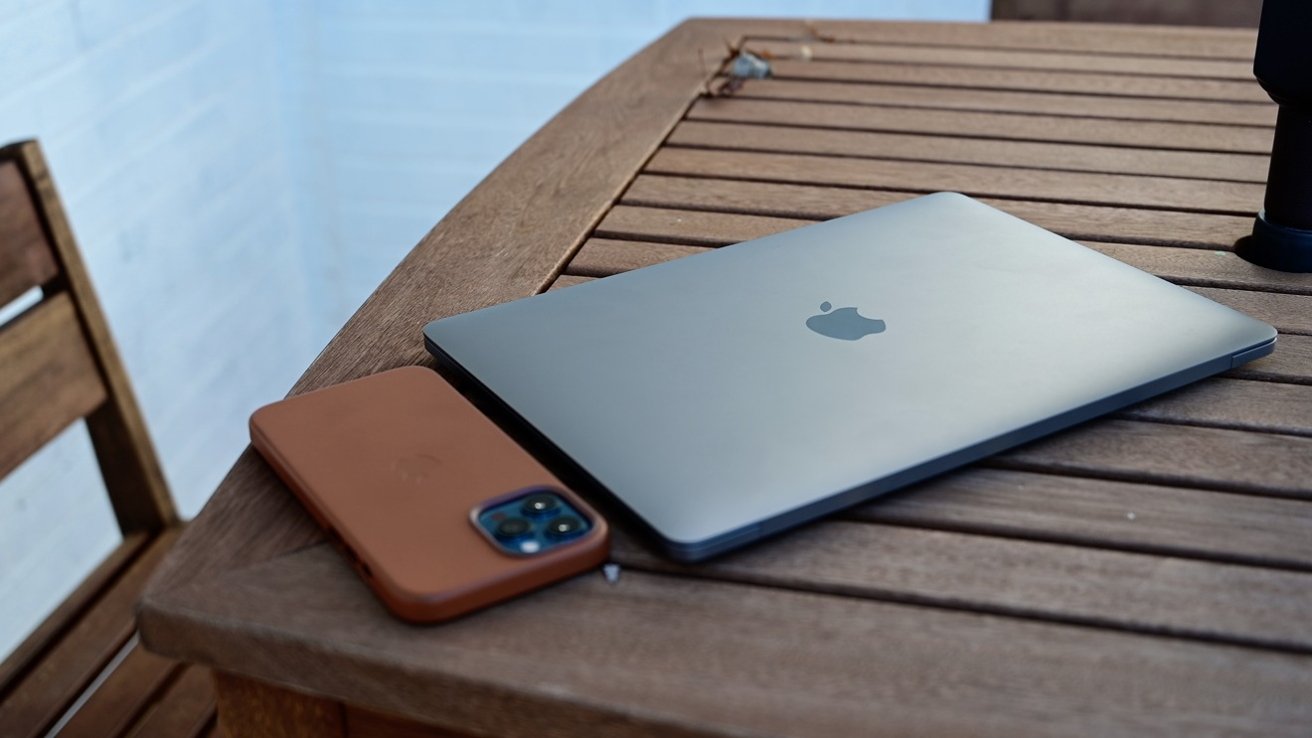 A closed MacBook and an iPhone in a brown case lie on a wooden table, next to a wooden chair.